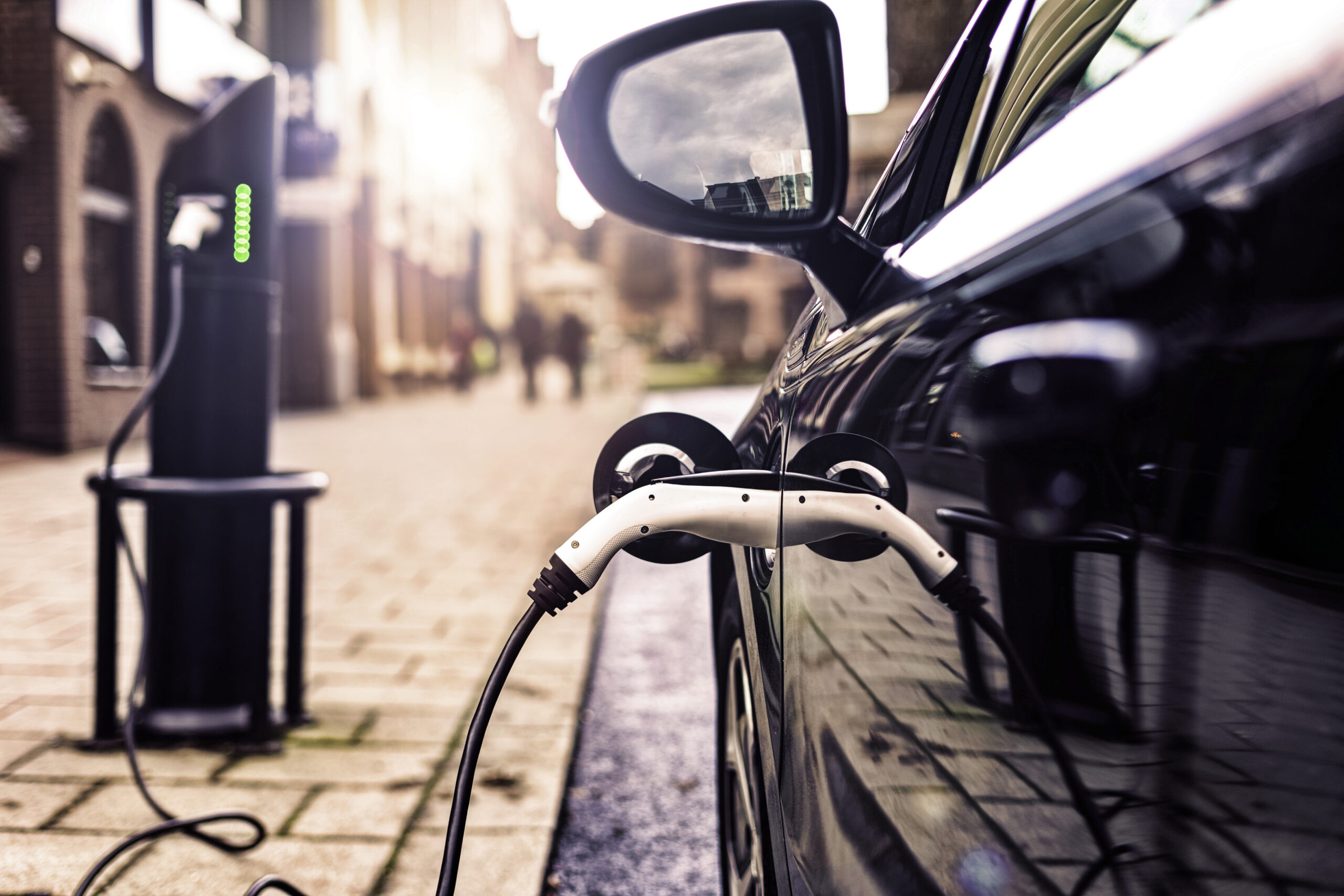 Close-up of an electric car parked on a city street, charging at a public charging station with the plug connected to the vehicle and buildings blurred in the background.