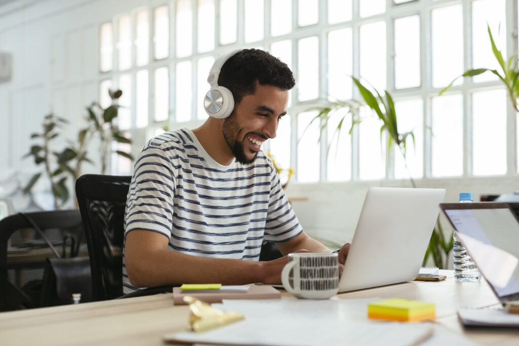 A man wearing headphones and a striped shirt smiles while using a laptop at a bright, modern desk with plants and office supplies around him.