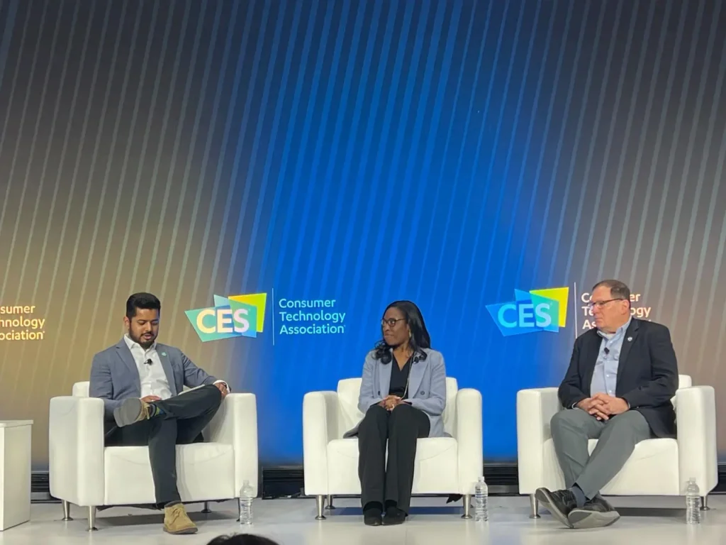 Three people sit on stage in white chairs during a CES event, with "Consumer Technology Association" logos displayed behind them. Two men and one woman appear to be participating in a panel discussion.