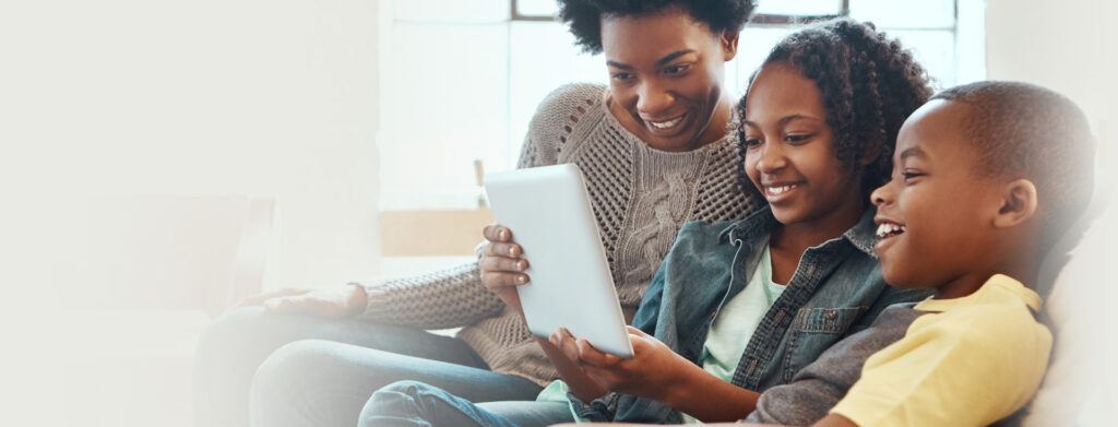 A woman and two children are sitting on a couch, smiling and looking at a tablet together in a bright, cozy room.