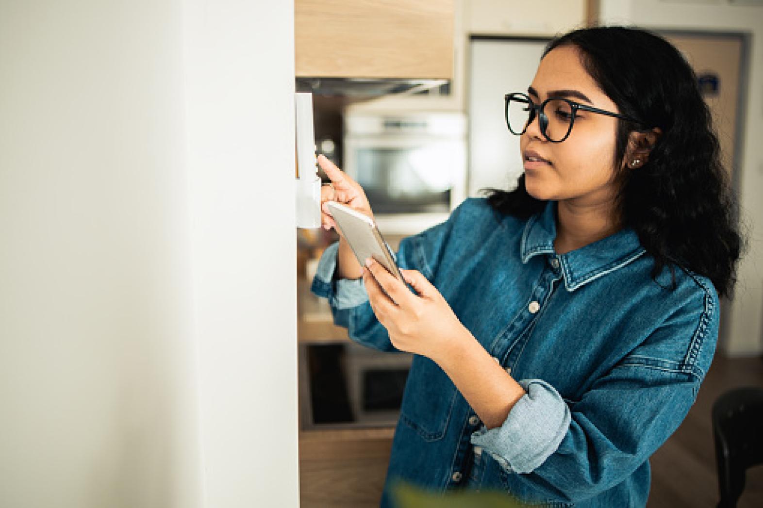 A woman with long curly hair and glasses uses her smartphone while adjusting a thermostat on the wall in a modern home.