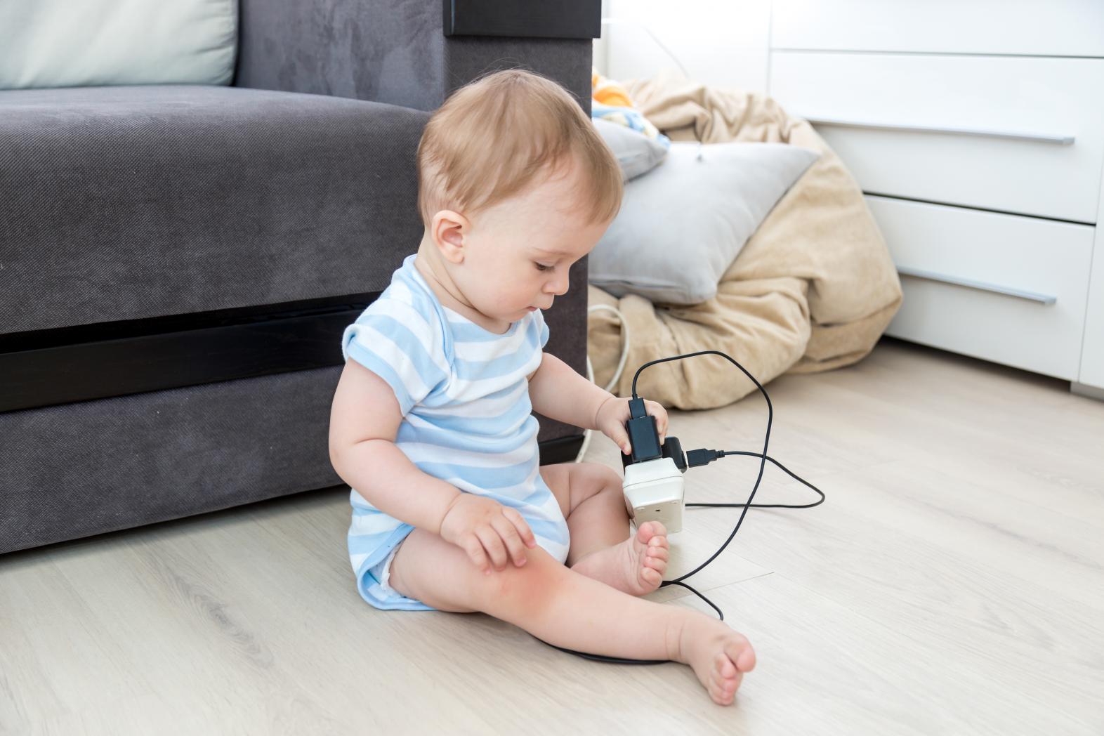 A baby in a striped onesie sits on the floor near a sofa, holding and playing with an electrical plug and socket, posing a safety hazard. The room is bright with household items in the background.