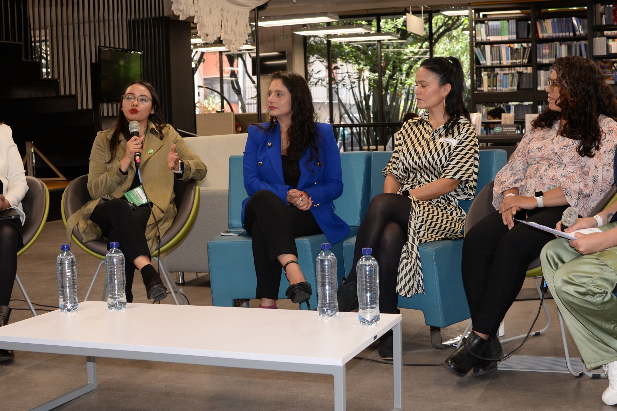 Five women sit in a semi-circle in a modern library, engaged in a panel discussion. One woman speaks into a microphone while others listen. Water bottles are on the table in front of them. Bookshelves and windows are in the background.