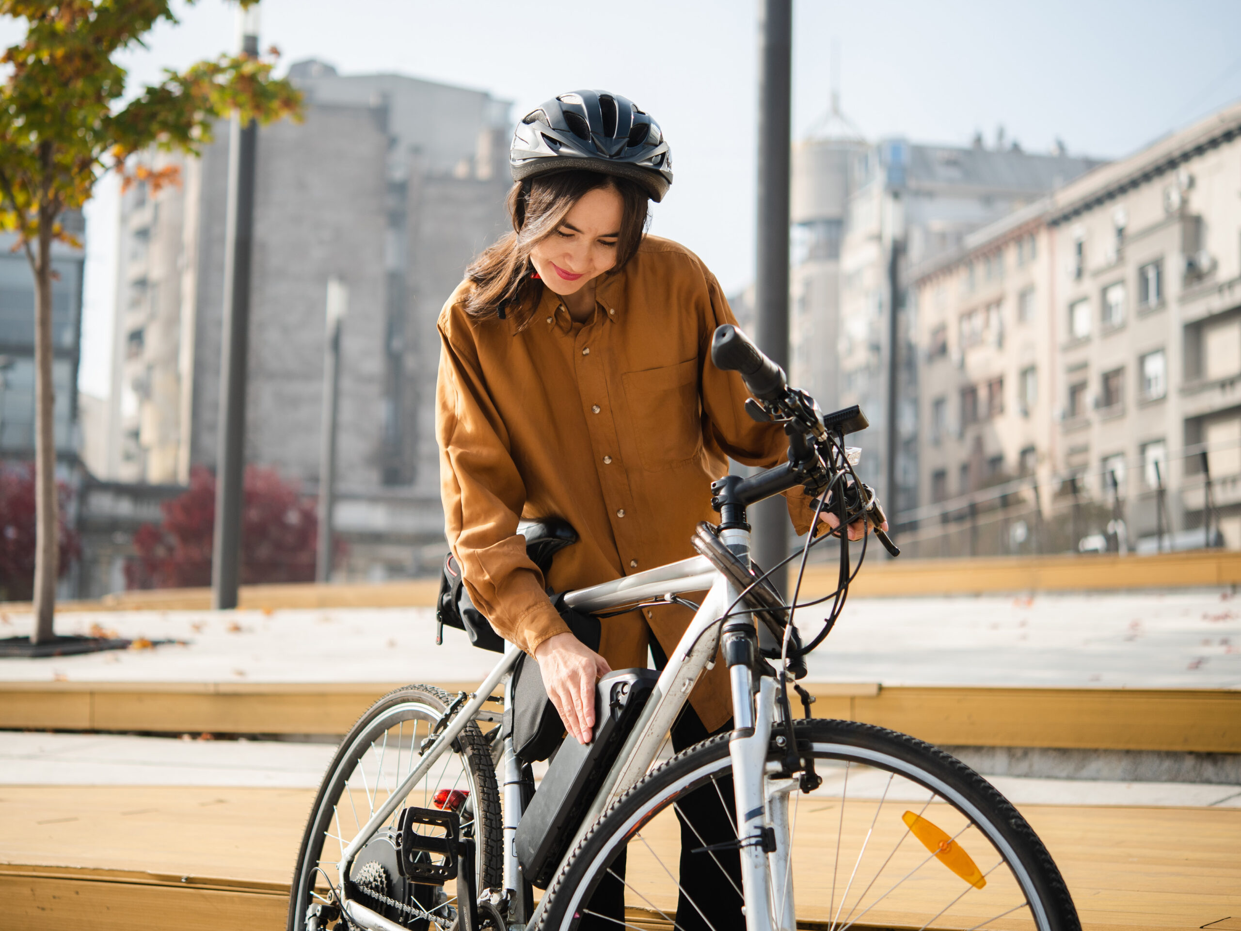 A woman in a brown shirt and black helmet stands outdoors, smiling while checking the battery on her electric bicycle. Urban buildings and trees are visible in the background.