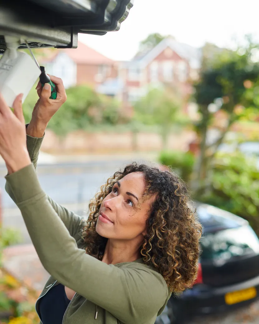 A woman with curly hair stands outside and installs or repairs a security camera under the eaves of a building, using a screwdriver. A parked car and houses are visible in the blurred background.