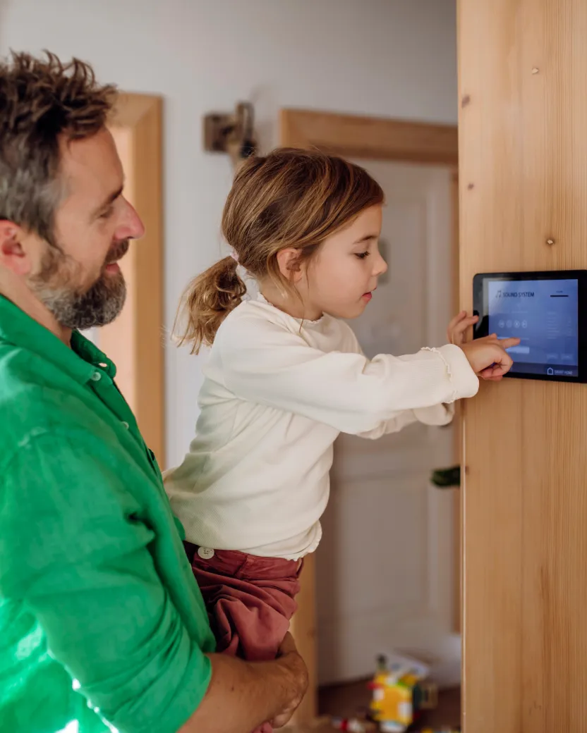 A man holds a young girl as she touches a smart home control panel mounted on a wooden wall, both focused on the screen. The setting appears to be a modern home interior.