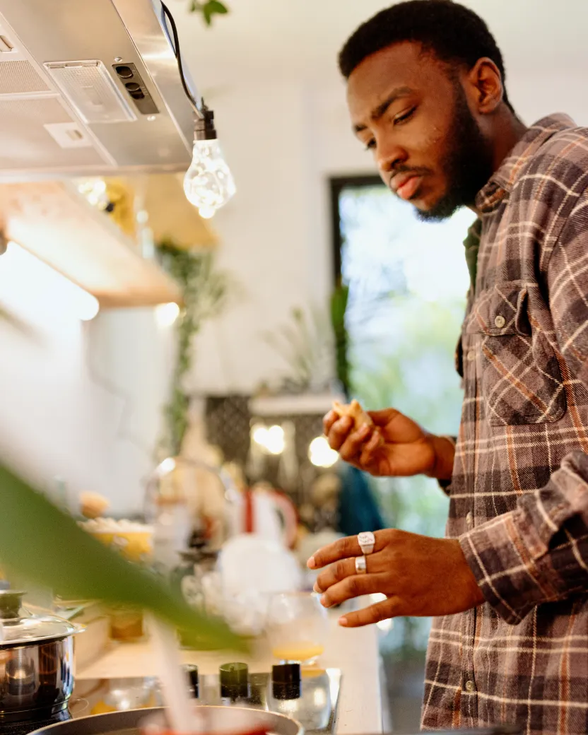 A man wearing a plaid shirt stands in a kitchen, holding food in one hand and tasting it. He appears focused on cooking, with pots on the stove and kitchen shelves in the background.
