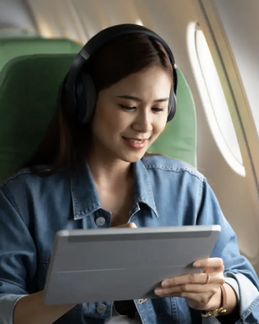 A woman wearing headphones and a denim jacket smiles while using a tablet on an airplane, seated next to a window.