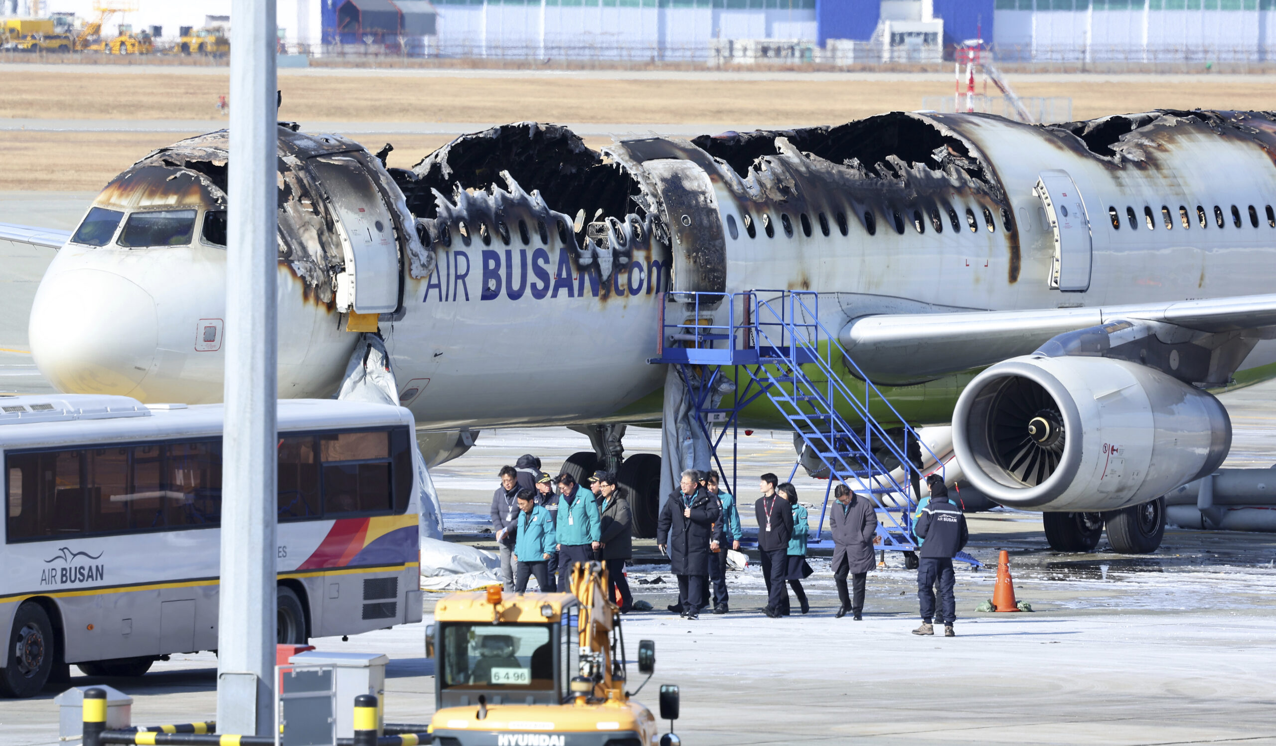 A severely burned Air Busan airplane with a large portion of its upper fuselage destroyed sits on the tarmac as investigators and personnel stand nearby, with an airport bus and vehicles in the foreground.