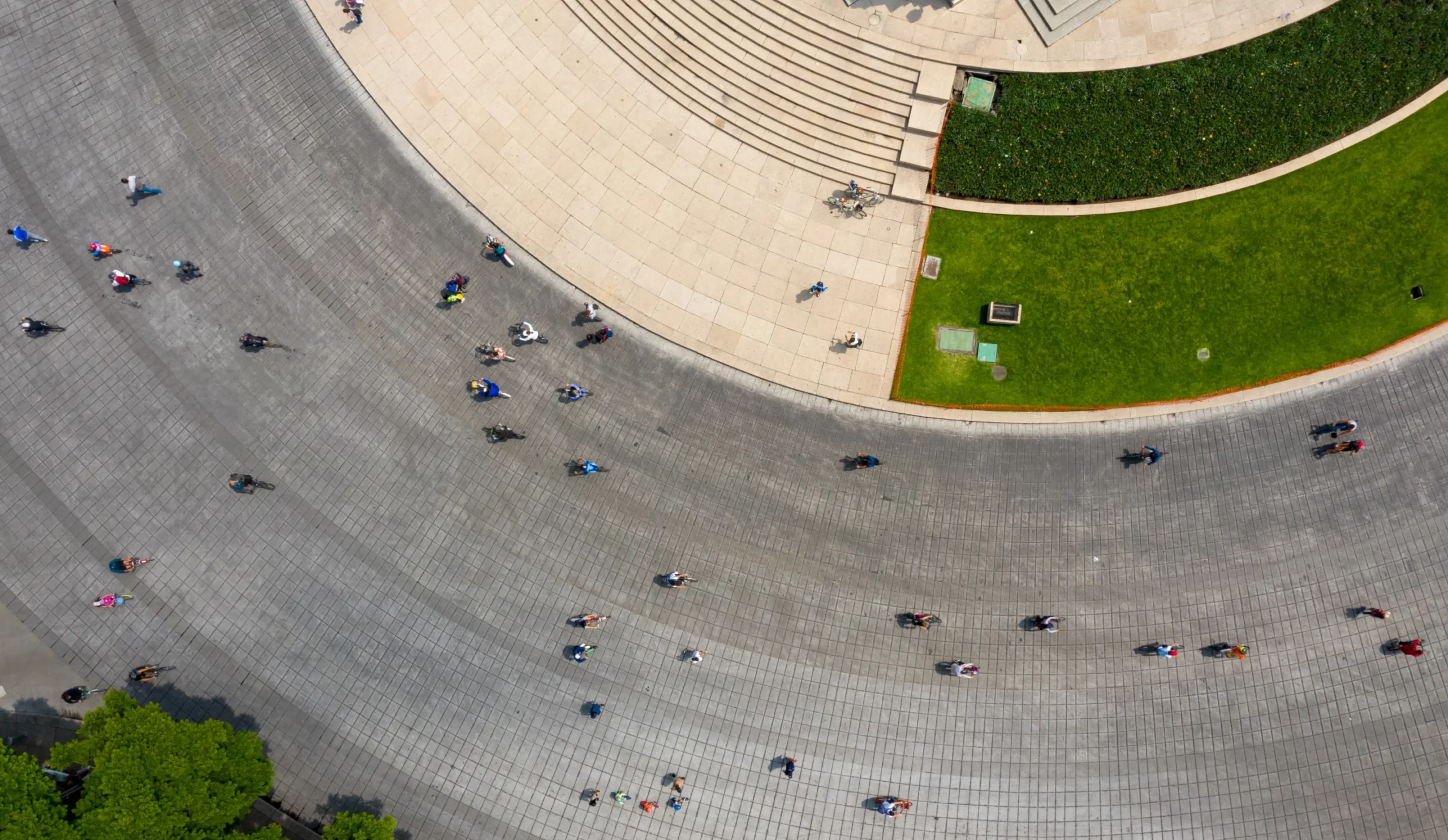 Aerial view of people walking on a wide, curved paved area near steps and green grass, with scattered shadows and some trees visible at the edges.