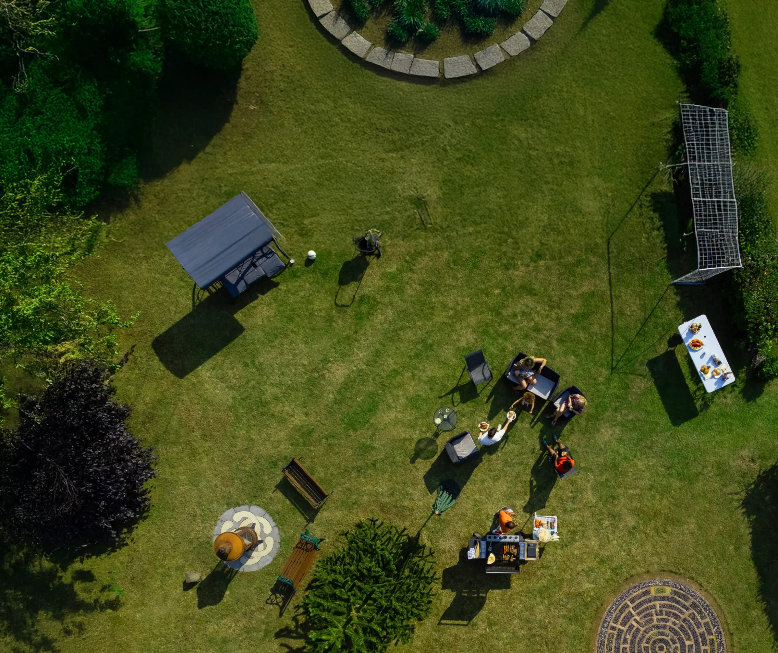 Aerial view of a group of people sitting around a table in a sunny garden, surrounded by greenery, benches, and a table with food. The scene is relaxed and summery.
