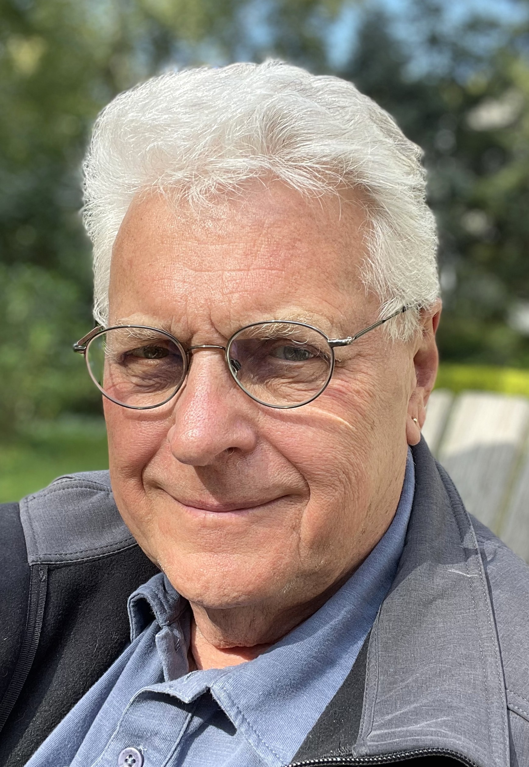 An older man with short white hair and glasses smiles slightly while sitting outdoors. He is wearing a blue collared shirt and a gray jacket, with greenery and sunlight in the blurred background.