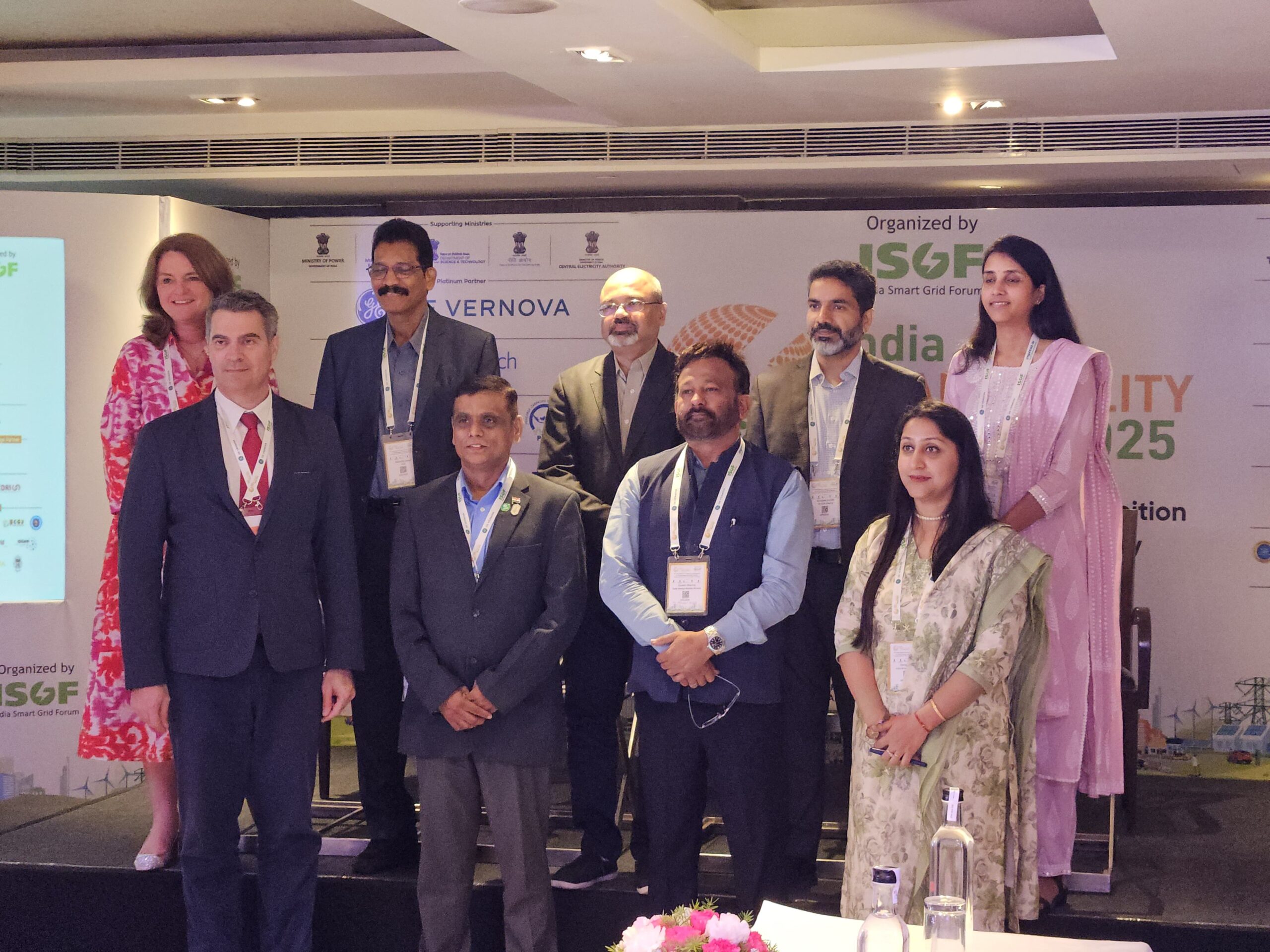 A group of ten people, both men and women in formal attire, stand and pose for a photo at an event. A backdrop displays logos and text about sustainability and India Smart Grid Forum.