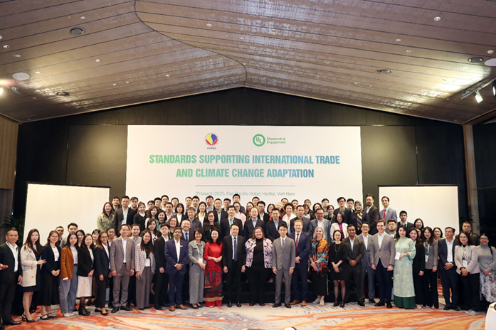 A large group of people in formal attire poses for a photo in front of a banner that reads "Standards Supporting International Trade and Climate Change Adaptation" at a conference event in a spacious hall.