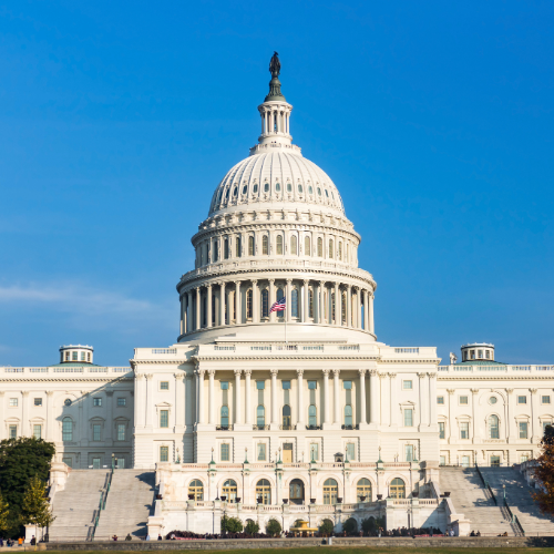 The United States Capitol building in Washington, D.C., with its iconic white dome, columns, and staircase, is shown against a clear blue sky. An American flag is flying above the entrance.
