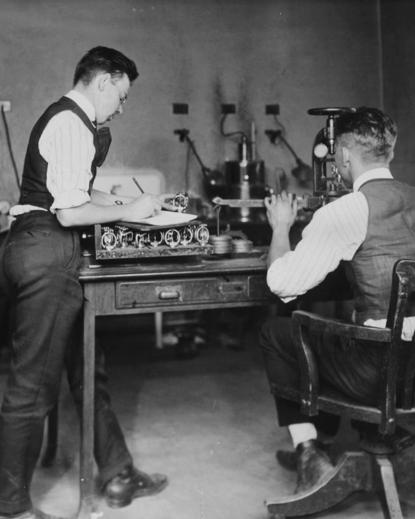 Two men in vintage clothing work in a laboratory; one is writing notes at a desk with scientific equipment, while the other sits and looks through a microscope. The setting appears to be from the early to mid-20th century.