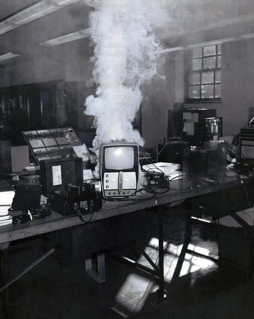 A black-and-white photo shows an old computer or monitor on a lab table, emitting a large cloud of smoke. The room is filled with other electronic equipment and sunlight streams through the windows.