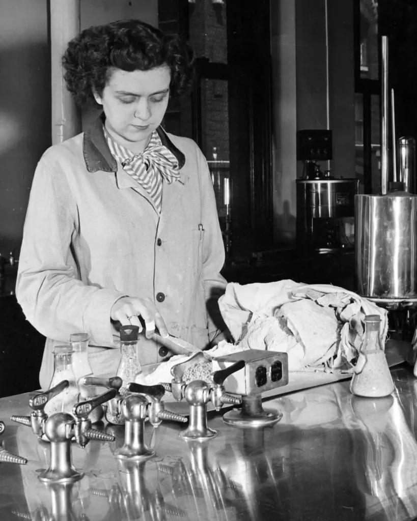 A woman in a lab coat works at a shiny laboratory table with various glassware, papers, and scientific equipment, appearing focused on her task.