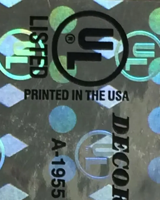 Close-up of a holographic sticker with the UL certification logo, "LISTED," "PRINTED IN THE USA," and "DECOR" along with the code "A-1955" on a patterned background.