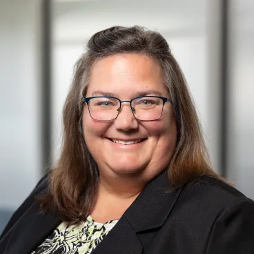A woman with long brown hair and glasses smiles at the camera. She is wearing a black blazer over a patterned top, and the background is softly blurred with light and vertical lines.