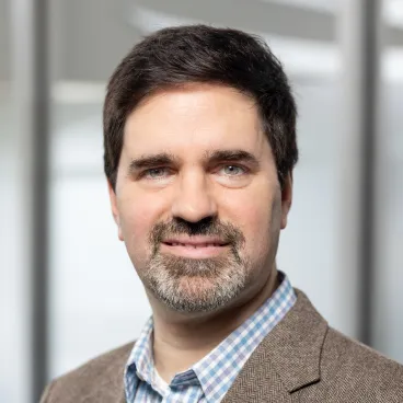 A man with short dark hair, a beard, and mustache, wearing a brown blazer over a blue and white checkered shirt, stands in front of a blurred background with light gray tones.