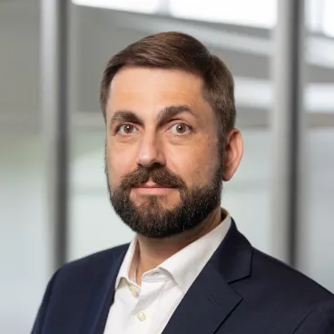 A man with short brown hair and a beard, wearing a dark suit jacket and white collared shirt, stands in front of large windows in a modern office setting.
