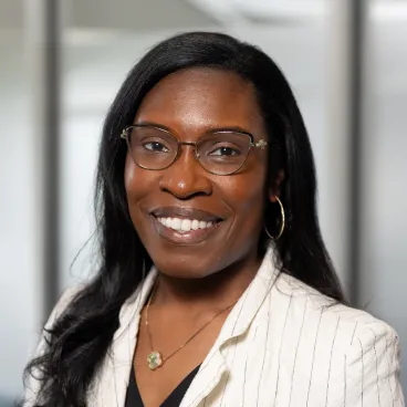 A woman with long black hair and glasses smiles, wearing a white pinstripe blazer, black top, gold hoop earrings, and a necklace, standing in an office setting with blurred windows in the background.