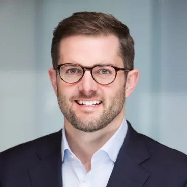 A man with short brown hair, a beard, and glasses is smiling. He is wearing a dark suit jacket and a light blue collared shirt. The background is softly blurred, likely an office setting.