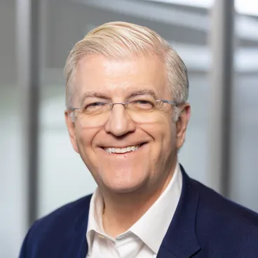 A smiling older man with light hair and glasses, wearing a dark suit jacket and white shirt, poses in front of a blurred office background.