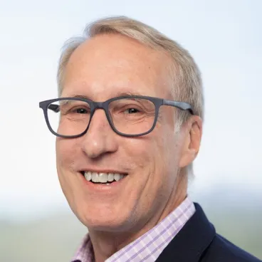 A smiling middle-aged man with short blond hair, wearing dark rectangular glasses, a checked shirt, and a dark blazer, posed in front of a blurred light background.