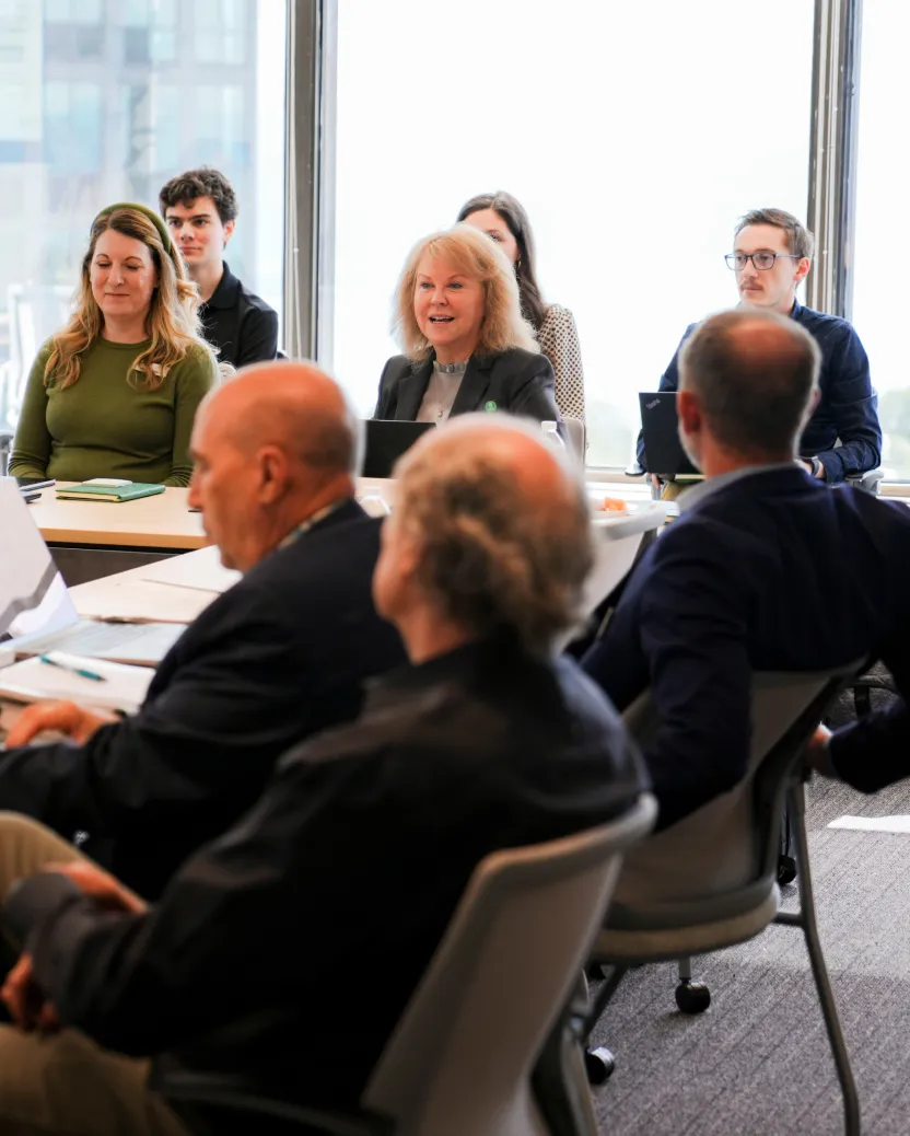 A group of people sit around a conference table in a modern office, engaged in a meeting. Some are listening attentively while one woman near the center is smiling and speaking. Large windows provide natural light.