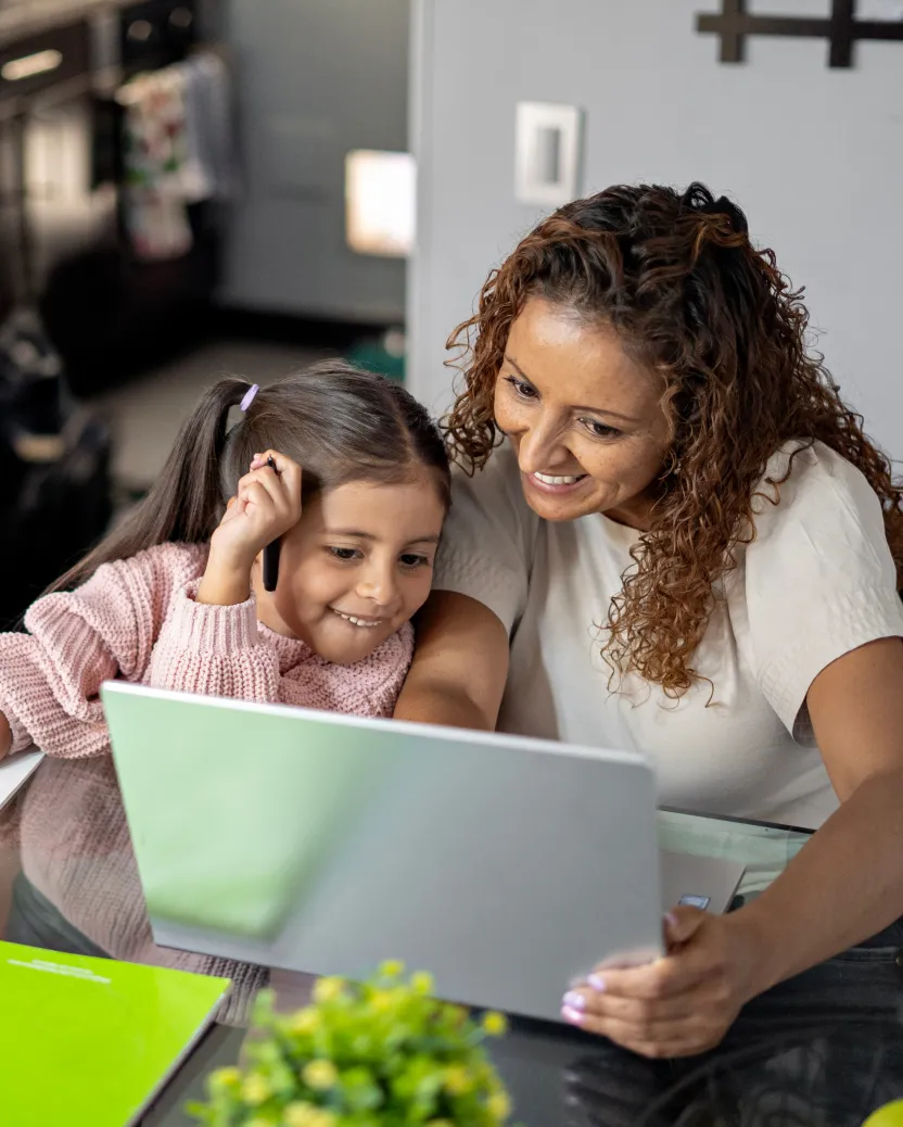 A woman and a young girl smile while looking at a laptop together at a kitchen table. The girl leans on the woman, and a green notebook and a small plant are visible on the table.