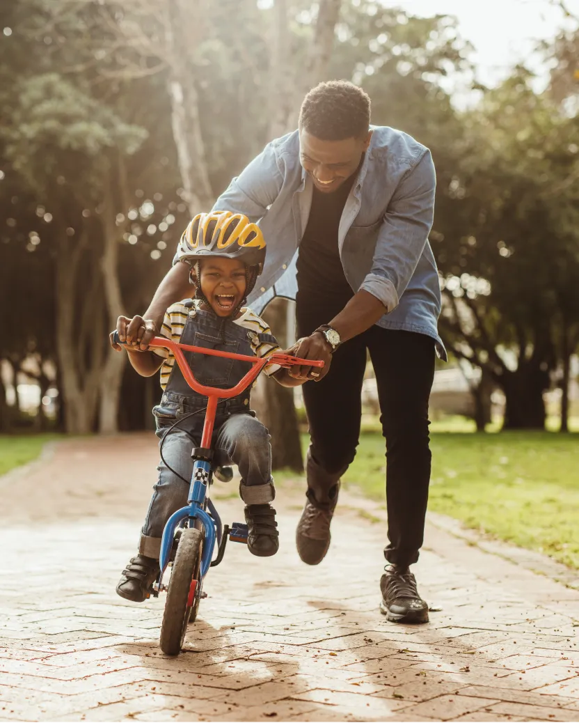 A smiling adult helps a laughing child ride a bicycle with training wheels on a park path. The child wears a yellow helmet and overalls, while the adult supports the bike, creating a joyful moment outdoors.