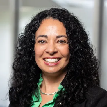 A woman with long, curly black hair smiles at the camera. She is wearing a green blouse, a black blazer, and a silver necklace. The background is softly blurred with natural light.