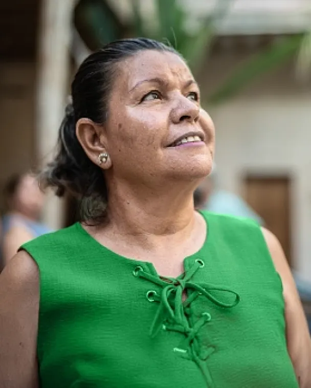 A woman wearing a green sleeveless top looks up and smiles, standing outdoors with blurred greenery and people in the background.