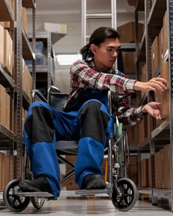 A person in a wheelchair, wearing a plaid shirt and blue work overalls, reaches for a box on a shelf in a warehouse aisle lined with cardboard boxes.