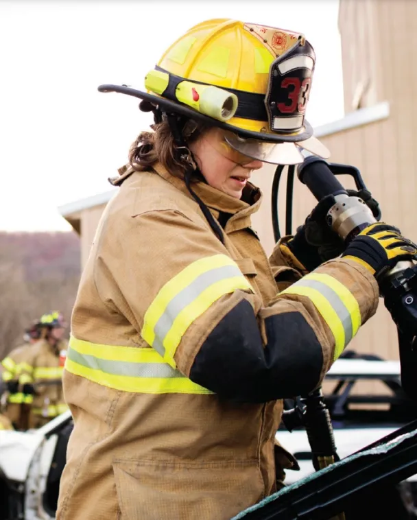 A firefighter in full gear, including a yellow helmet and reflective jacket, operates rescue equipment outdoors near a damaged vehicle. Another firefighter is visible in the background.