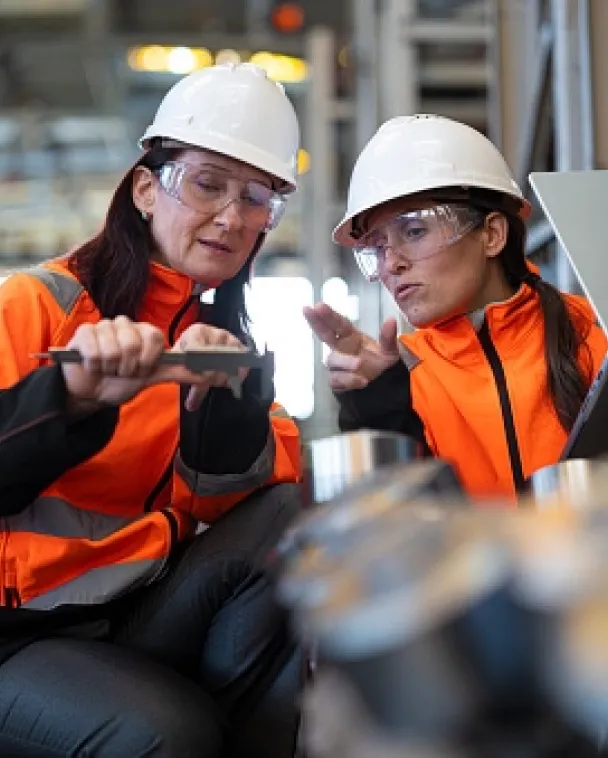 Two women wearing hard hats, safety glasses, and orange jackets are examining a tool and discussing in an industrial setting, with machinery and blurred objects in the foreground.