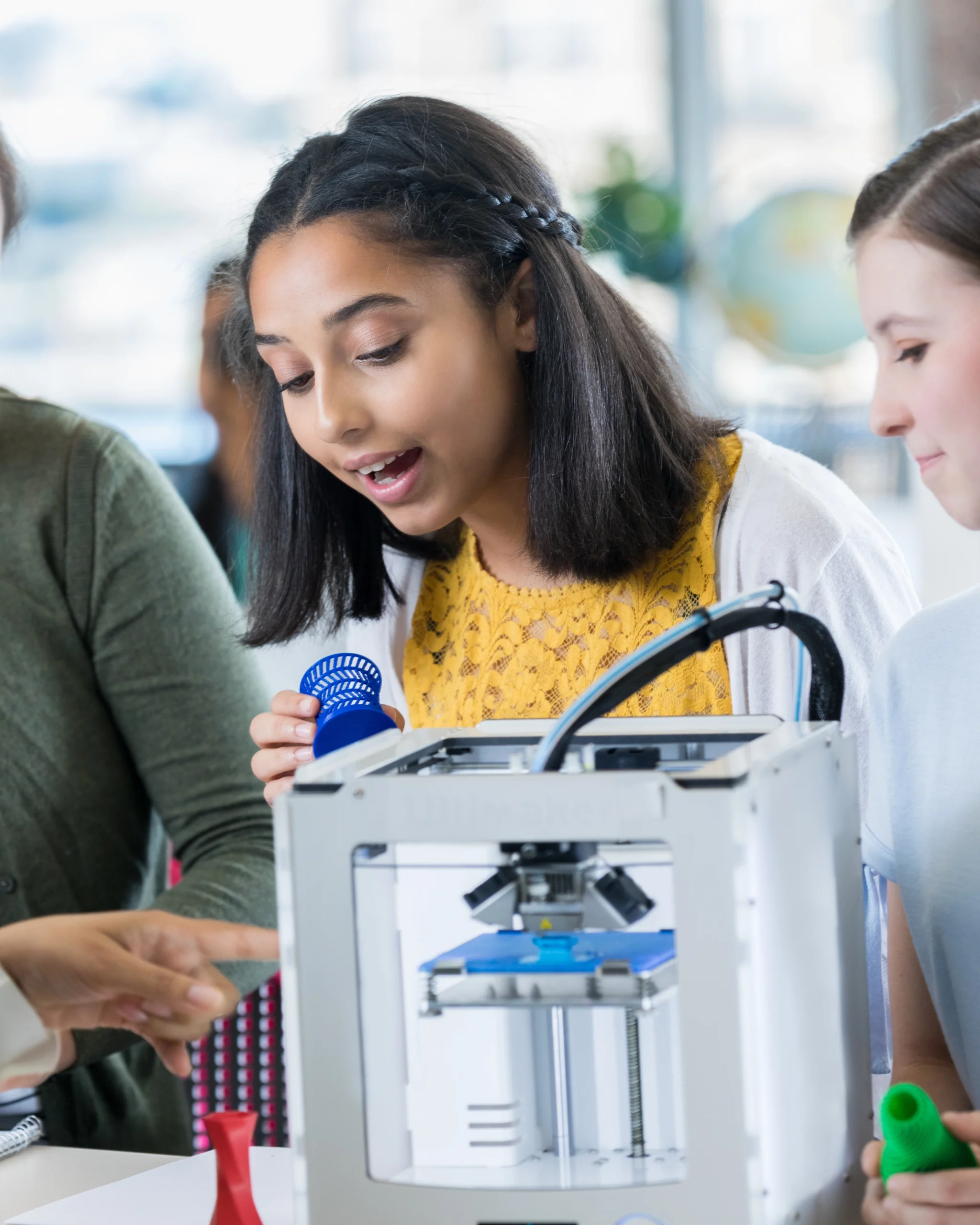 A young girl looks amazed while holding a 3D-printed object next to a 3D printer, surrounded by classmates and an adult in a bright classroom setting.