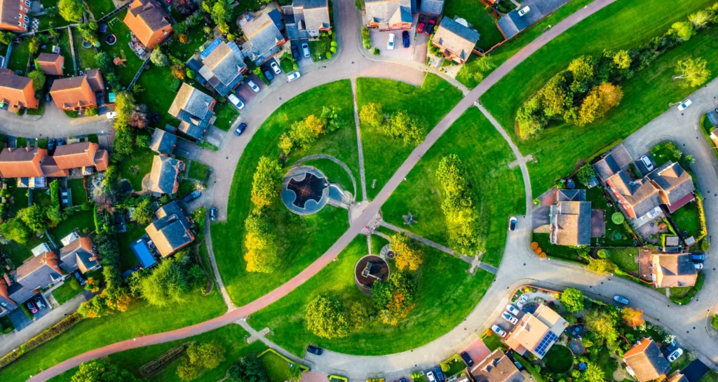 Aerial view of a circular park with green lawns, walking paths, and trees, surrounded by houses with red roofs, driveways, and parked cars in a suburban neighborhood.
