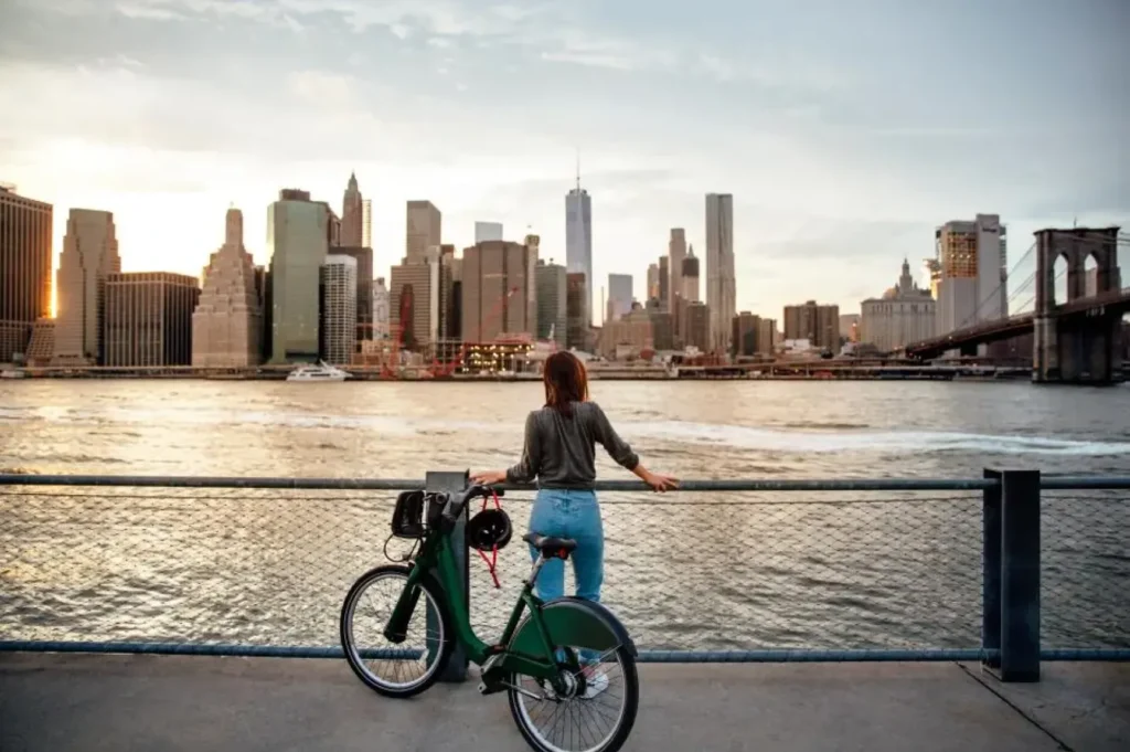 a woman standing on a bridge with a bicycle and a city in the background