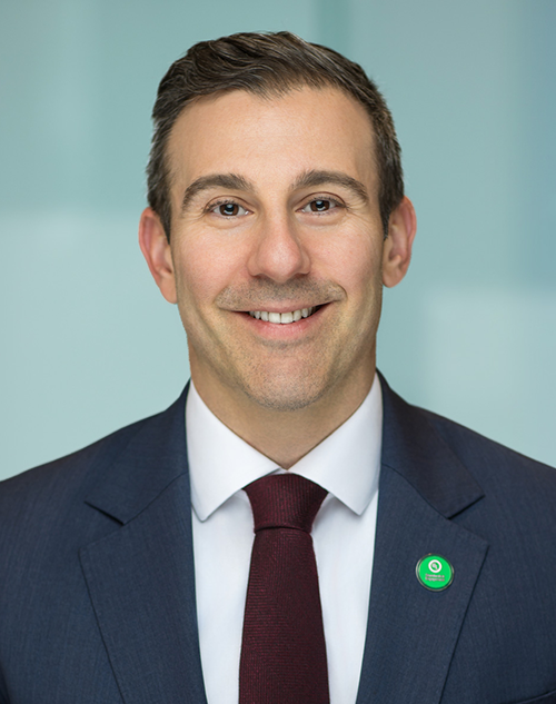 Smiling man in a dark suit, white shirt, and burgundy tie, wearing a green circular pin on his lapel, standing in front of a light blue blurred background.