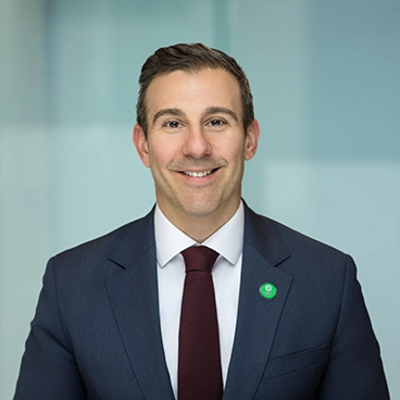A man in a dark suit, white shirt, and maroon tie is smiling at the camera. He is wearing a green pin on his lapel. The background is blurred and light-colored.