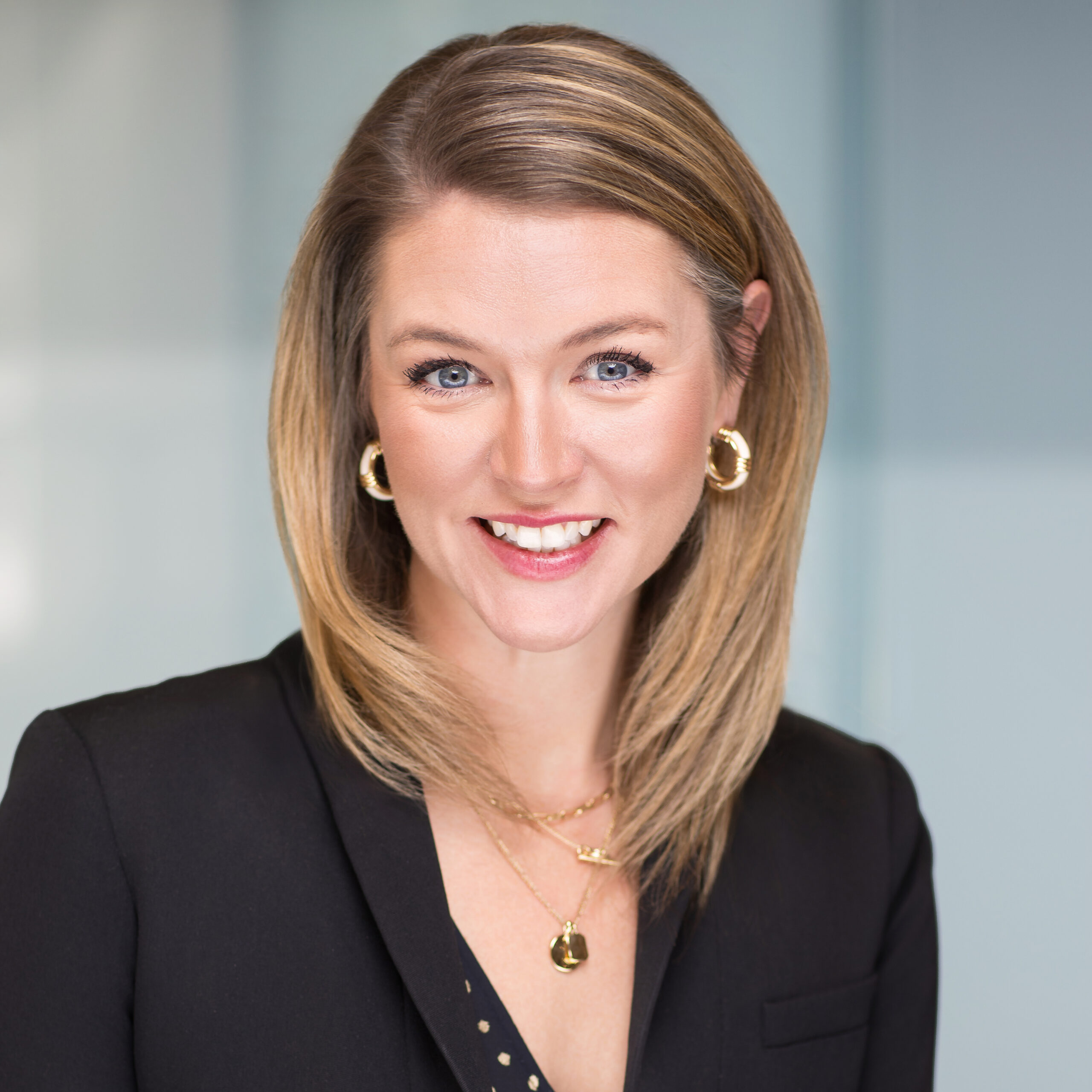 A woman with straight, shoulder-length blonde hair smiles at the camera. She is wearing a black blazer, gold hoop earrings, and layered gold necklaces. The background is blurred and softly lit.