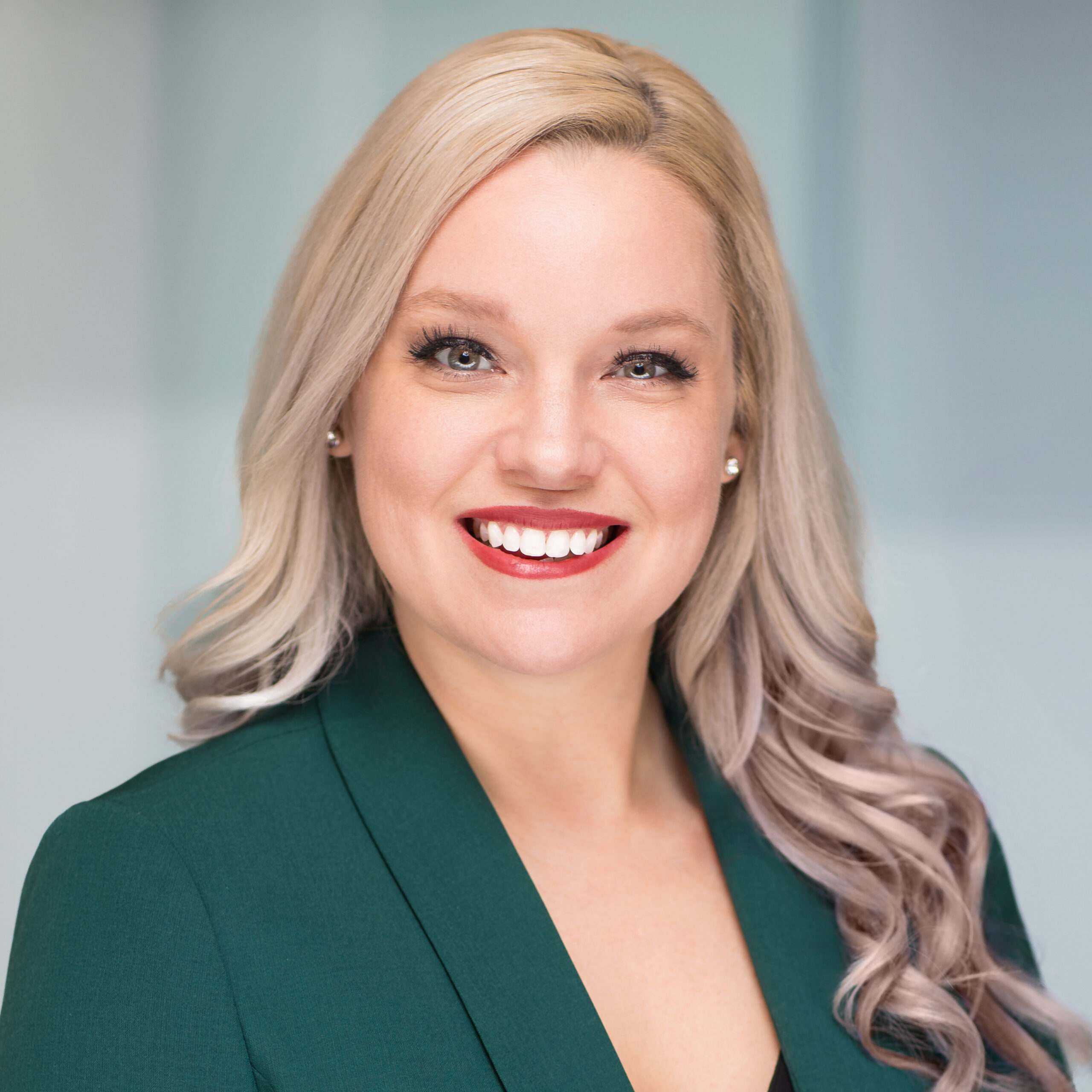 A woman with long, wavy blonde hair smiles at the camera. She is wearing a dark green blazer over a black top, with a light, blurred background.