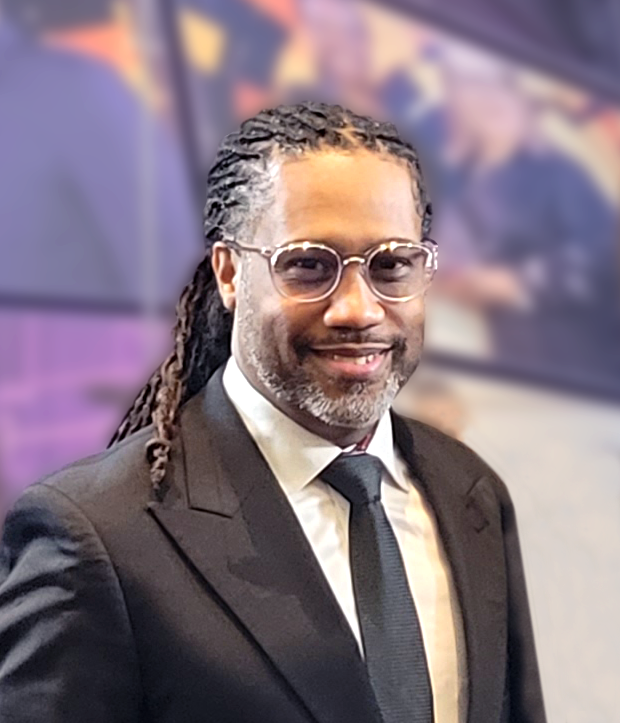 A man with long, braided hair wearing glasses, a black suit, white shirt, and black tie, smiles at the camera. The background is blurred with purple and yellow tones.