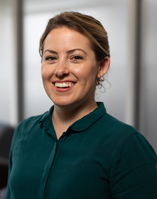 A woman with light skin and brown hair pulled back, wearing a dark green collared shirt, smiles at the camera in a softly lit indoor setting with blurred background.
