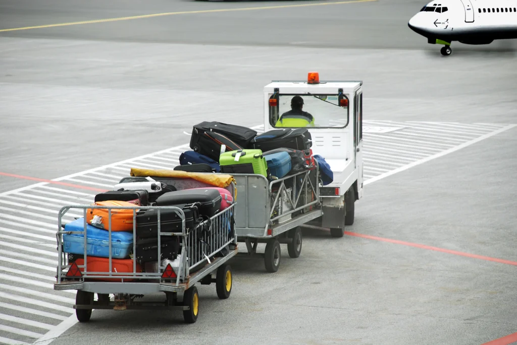 A baggage cart loaded with colorful suitcases is being towed by a small vehicle on an airport tarmac, with part of an airplane visible in the background.