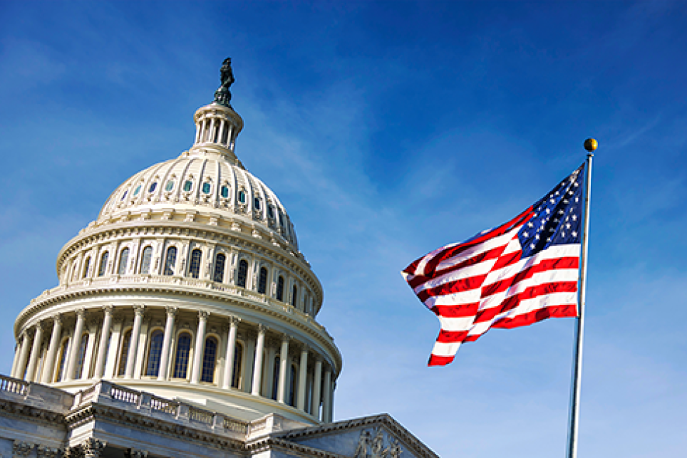 The U.S. Capitol building’s dome and a waving American flag are shown under a clear blue sky.