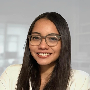 A young woman with long, straight dark hair and clear glasses smiles at the camera. She is wearing a light-colored top, and the background is a softly blurred indoor office setting.